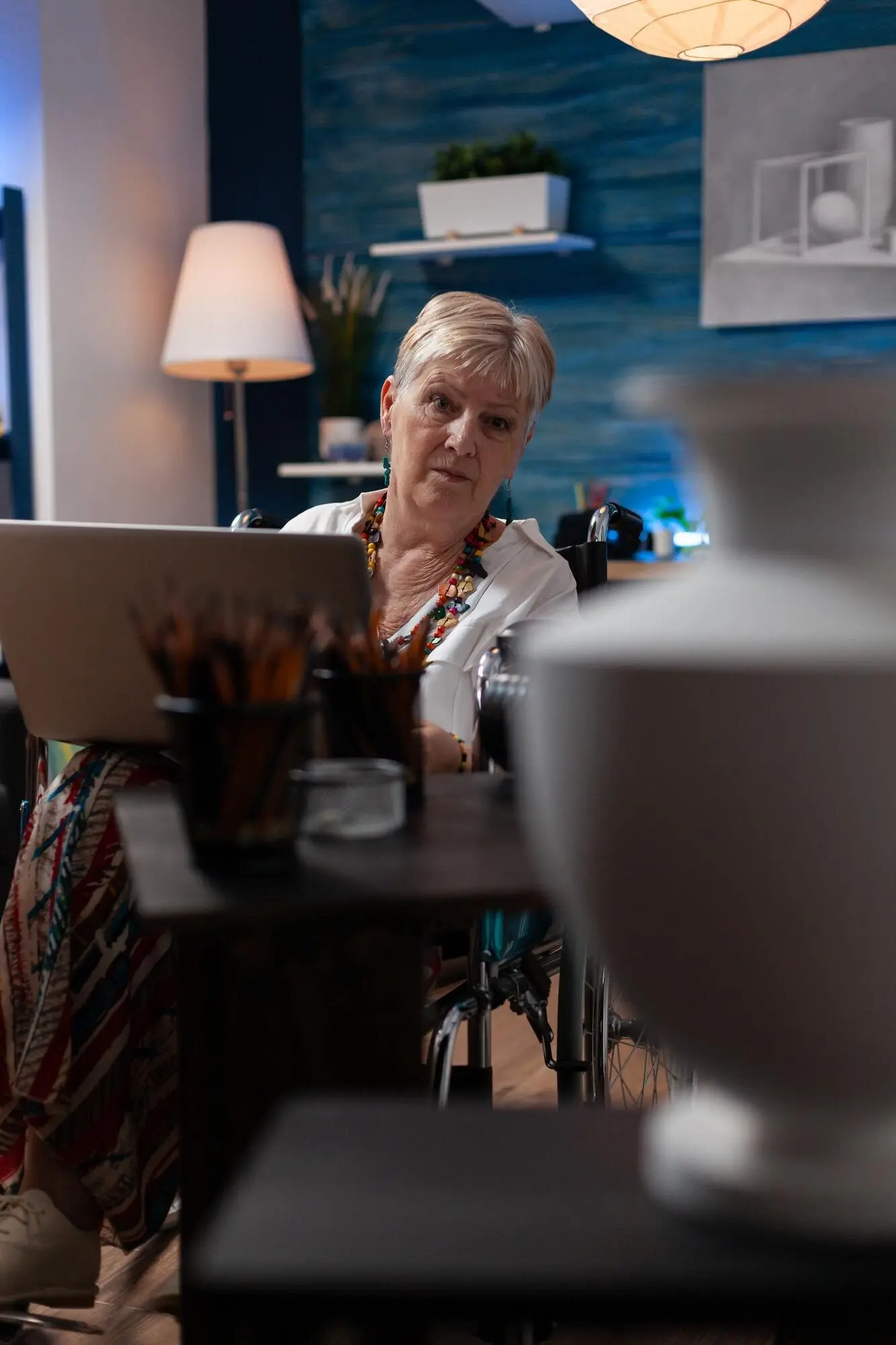 Пожилая женщина‑художница, живущая с инвалидностью, в домашней художественной студии использует ноутбук и смотрит на вазу в поисках вдохновения. Она сидит в инвалидной коляске, держит портативный компьютер и просматривает идеи для набросков.